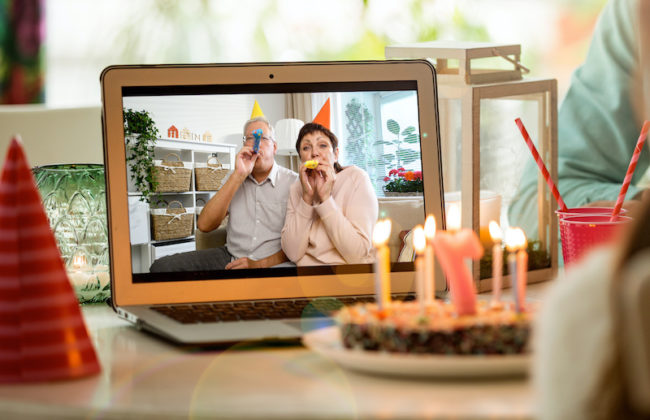 Happy little girl celebrating birthday at home with parents and grand parents on video call. Laptop with senior couple online, cake with candles on table.